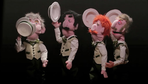 A puppet barber shop quartett performing. They are all holding hats in their hands, one of the hats has a bite taken out of it.