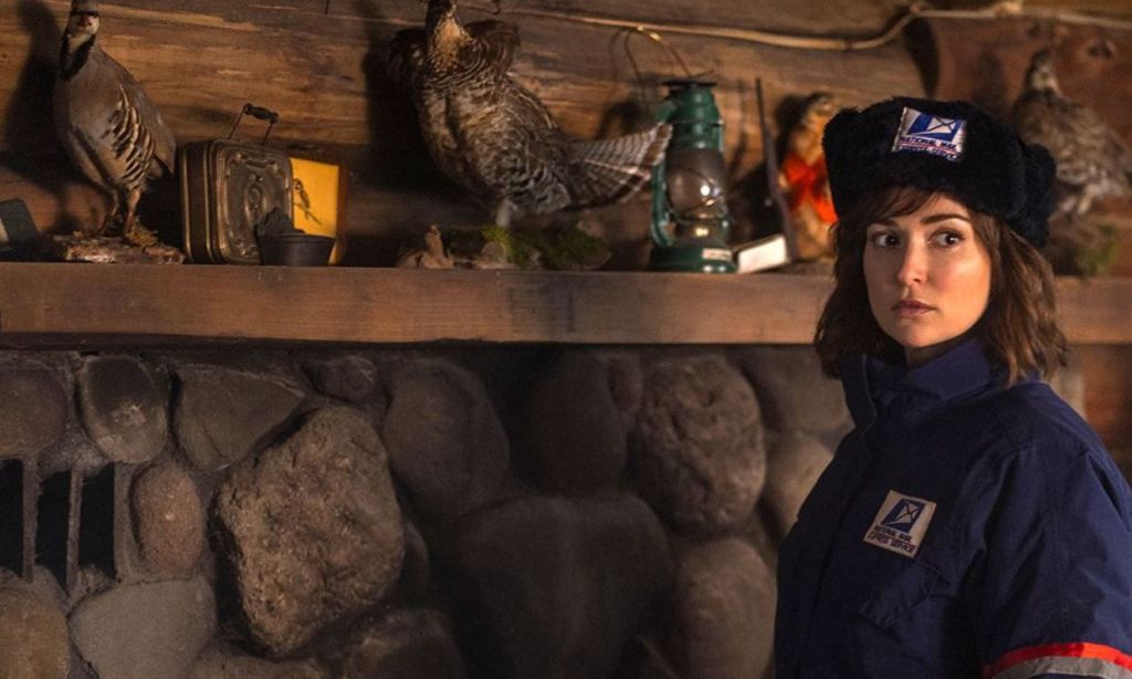 Cecily (Milana Vayntrub) standing in front of a fireplace in her USPS uniform.