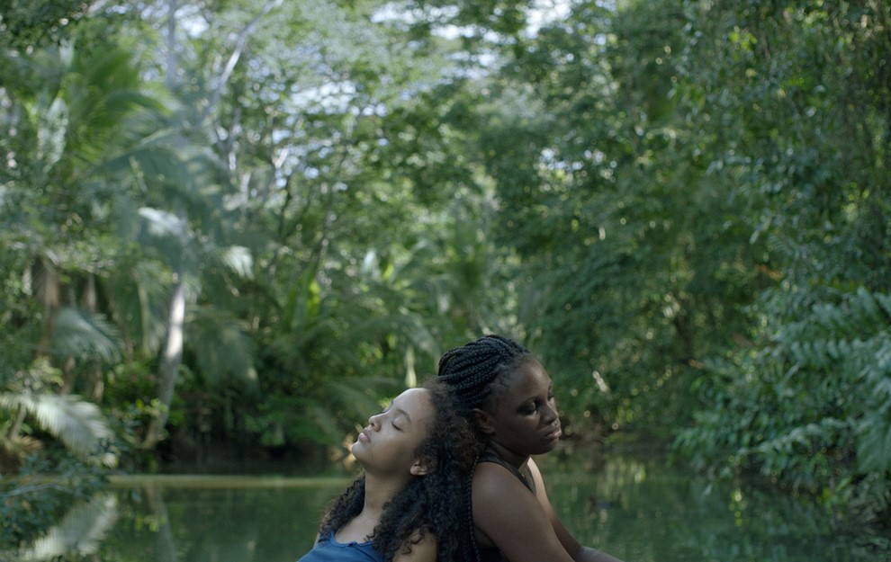 Selva (Smashleen Gutiérrez) sitting back to back with Winter (Keha Brown) in the forest next to the river.
