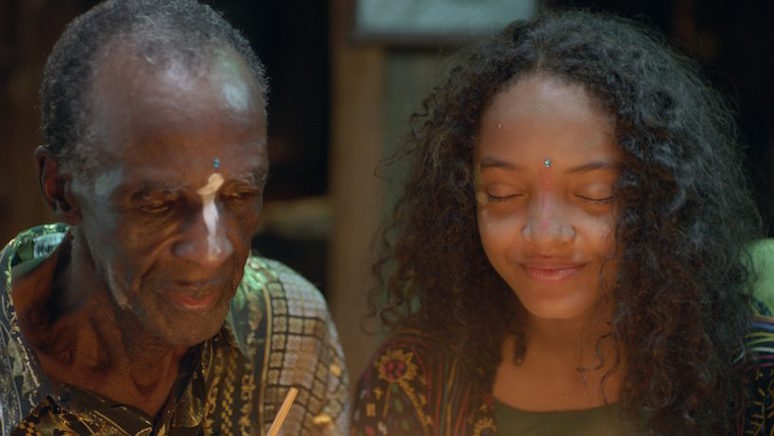 Selva (Smashleen Gutiérrez) and her grandfather (Humberto Samuels) with their eyes closed and a bindi-like stone on their foreheads, and color on their noses.
