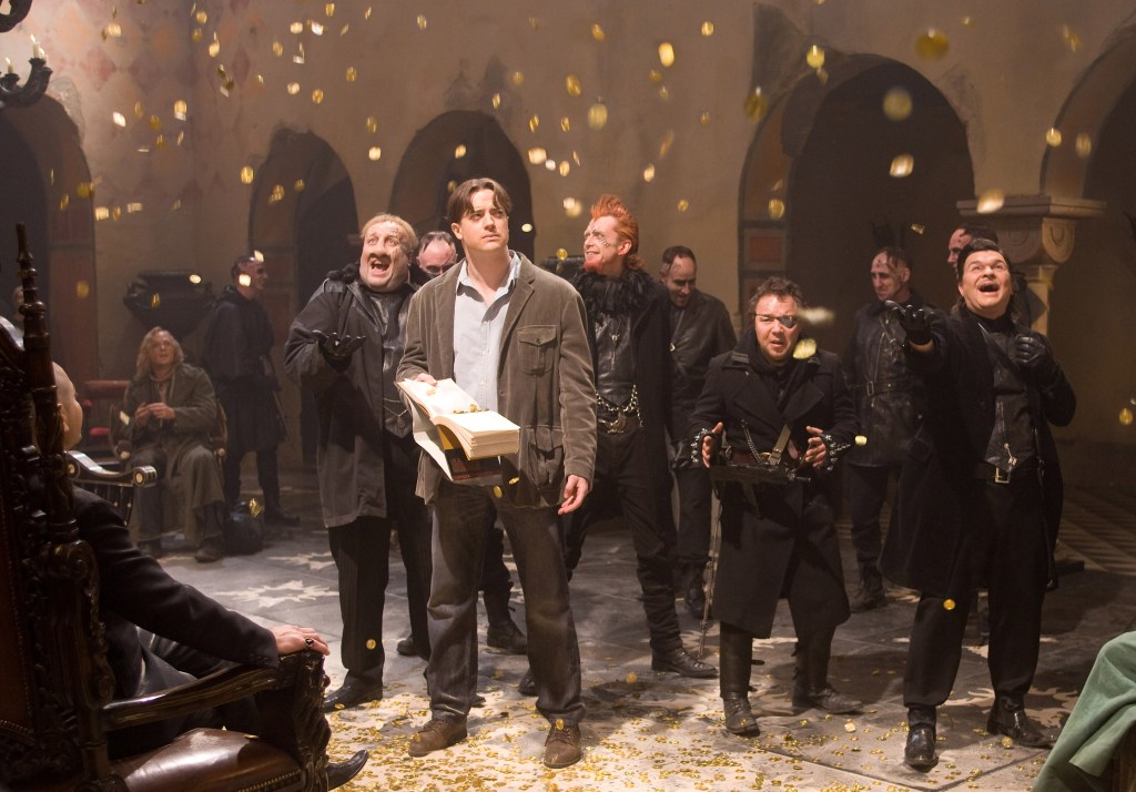 Mo (Brendan Fraser) holding an opne book, surrounded by Capricorn (Andy Serkis) and his men as golden coins rain down on them.