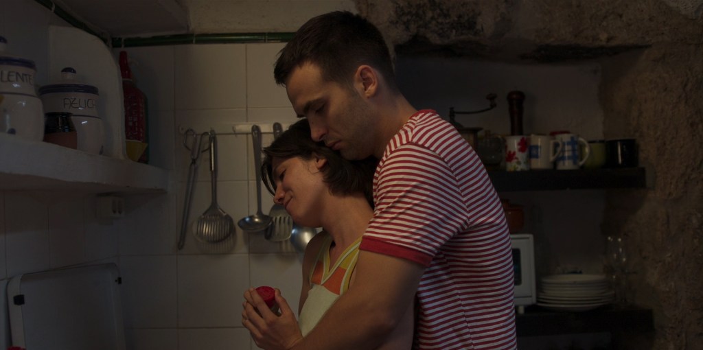 Moi (Ricardo Gómez) hugging Mía (Bruna Cusí) from behind in the kitchen.