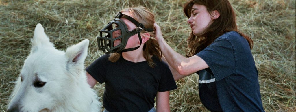 Gloria (Alba Gaïa Bellugi) putting a dog muzzle on Lucie (Janaina Halloy) as they play with the dog.