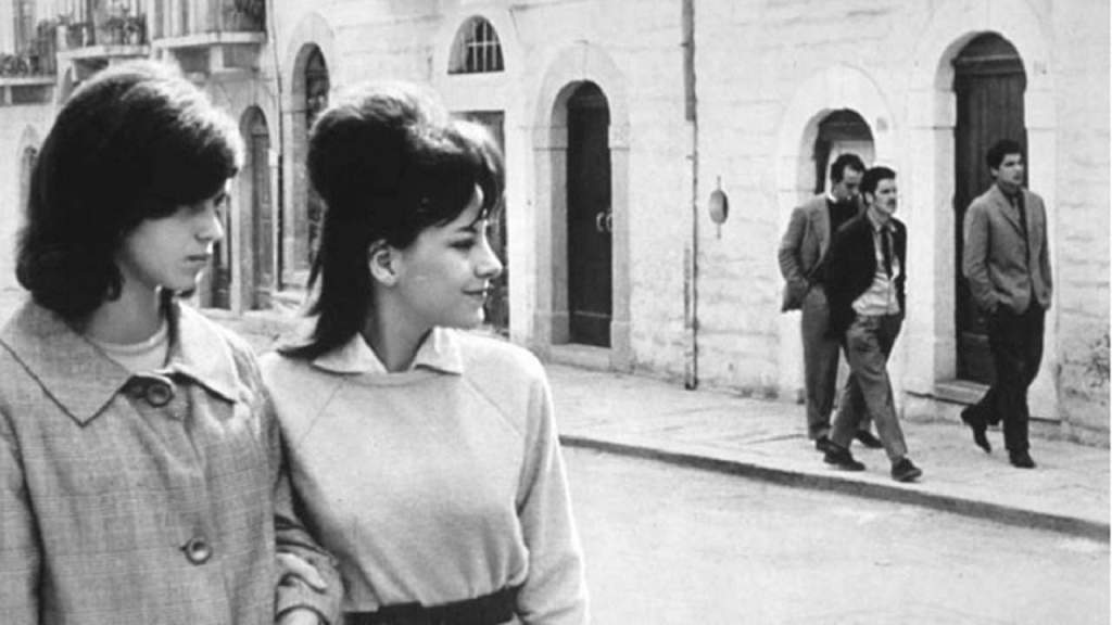 Two young women walking arm in arm, looking at the three guys on the other side of the street.