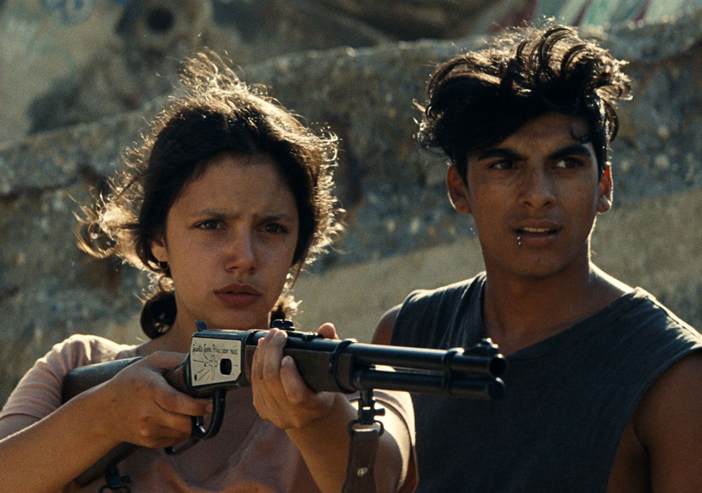 Ava (Noée Abita) aiming a rifle, with Juan (Juan Cano) standing next to her.