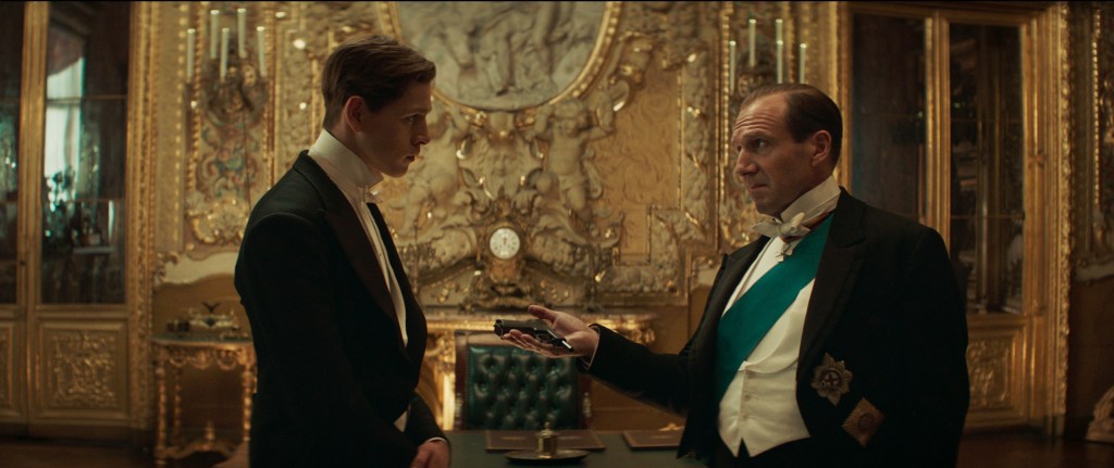 Orlando (Ralph Fiennes) giving Conrad (Harris Dickinson) a gun. 