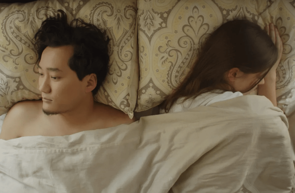 Anar (Iveel Mashbat) and Kassy (Jana Miley) lying in bed, turned away from each other.