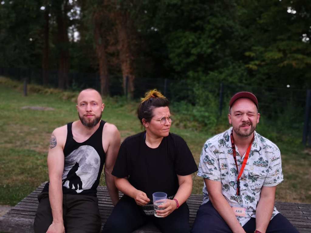 The band sitting on a picnic table in a park - two men and a woman.