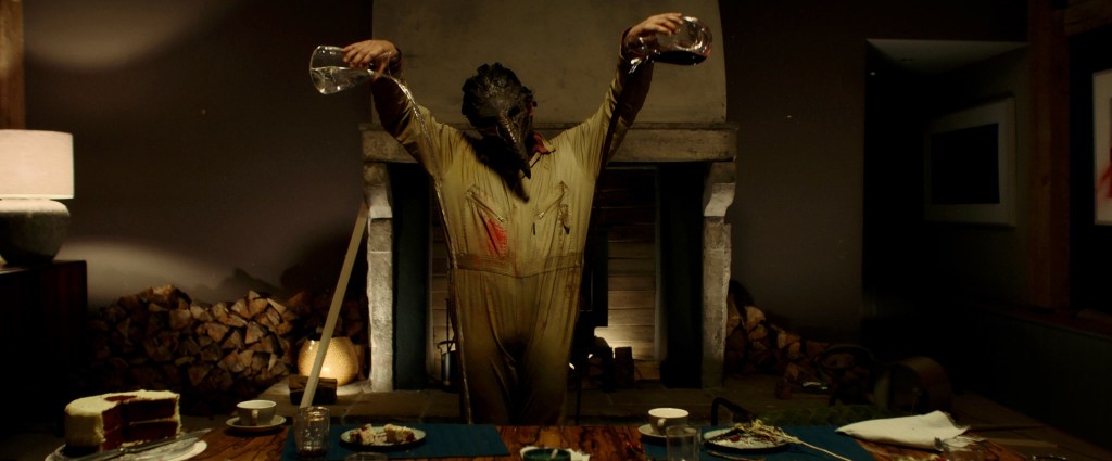 A man in a bird mask and an overall pouring water and wine over the dinner table.