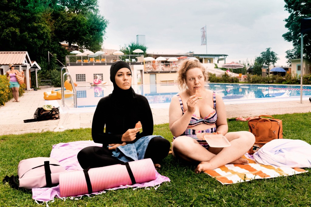 Yasemin (Nilam Farooq), wearing a burkini, and Paula (Julia Jendroßek), wearing a bikini, sitting next to the pool together.