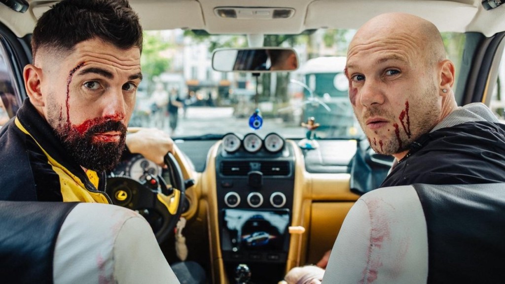 Noah (Dimitri 'Vegas' Thivaios) and Carlos (Jeroen Perceval) sitting in the front seats of the car, looking back. They both have blood on their faces.