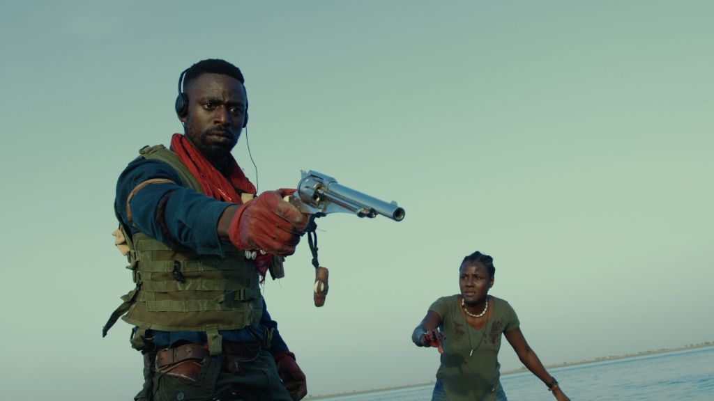 Chaka (Yann Gael) aiming a revolver at something. Awa (Evelyne Ily Juhen) stands behind him.