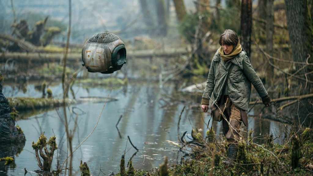 Vesper (Raffiella Chapman) walking through the forest accompanied by a cubic drone with a worried face painted on one side..