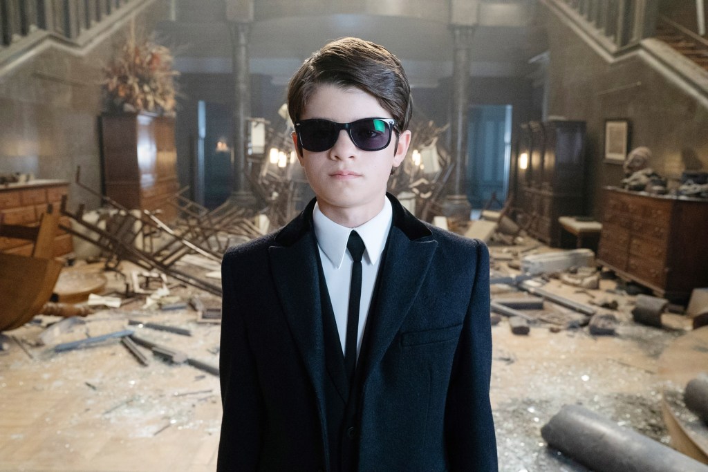 Artemis Fowl (Ferdia Shaw) wearing sunglasses, standing in a completely messed up entry hall. 