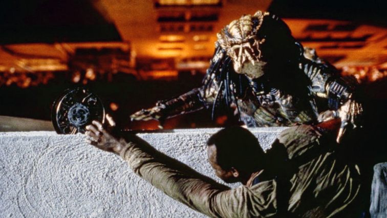 The Predator (Kevin Peter Hall) hanging off a building ledge, holding on to Mike Harrigan (Danny Glover) who is reaching for an explosive device lodged in the wall.