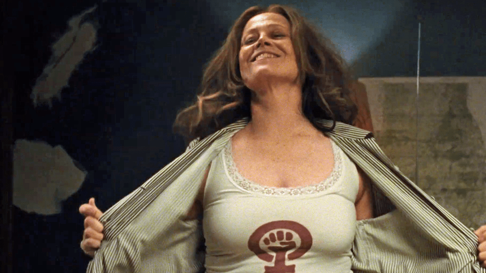 Virginia (Sigourney Weaver) pulling open her blouse. Below it, she is wearing a tank top with a feminist symbol.