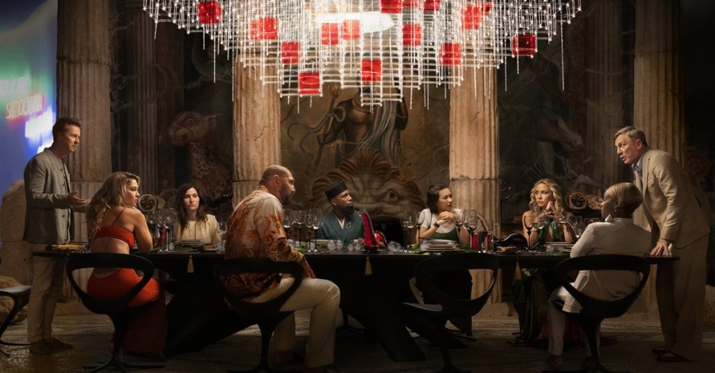 Miles Bron (Edward Norton) confronted by Benoit Blanc (Daniel Craig) across a long table at which the entire group is sitting. 