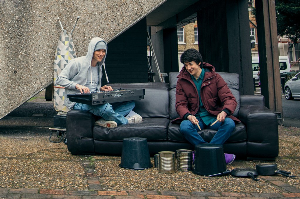Vince (Ed Skrein) and Stevie (Leo Long) sitting on a couch in front of Stevie's apartment building. Vince is playing on a keyboard and Stevie is drumming on different bins and cans.