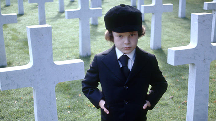 Damien (Harvey Stephens) standing in a military cemetery between the graves.