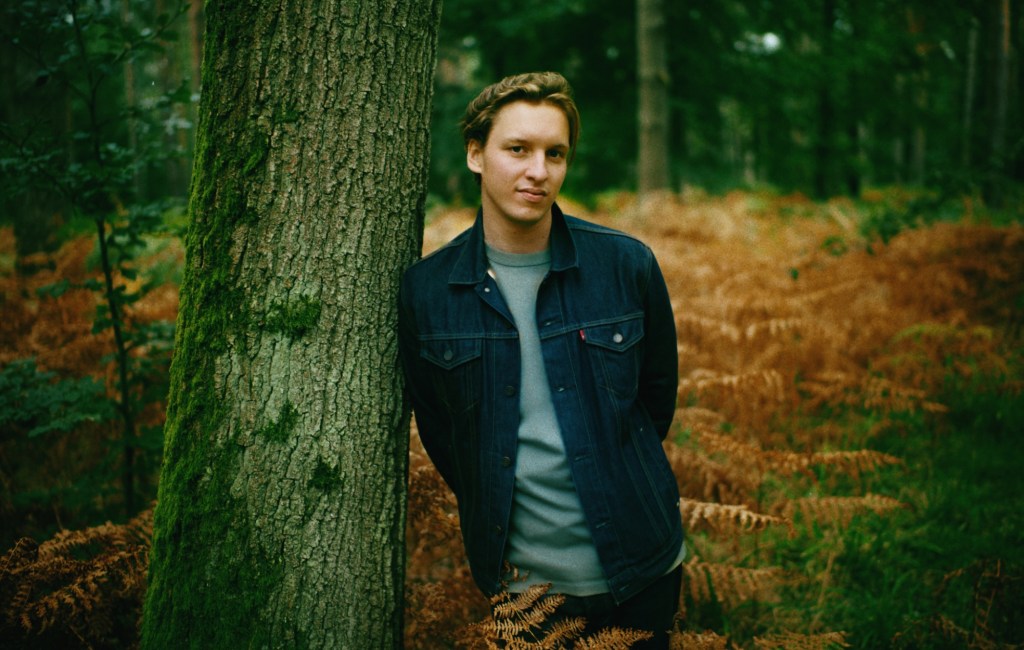 George Ezra leaning against a tree in a forest. 