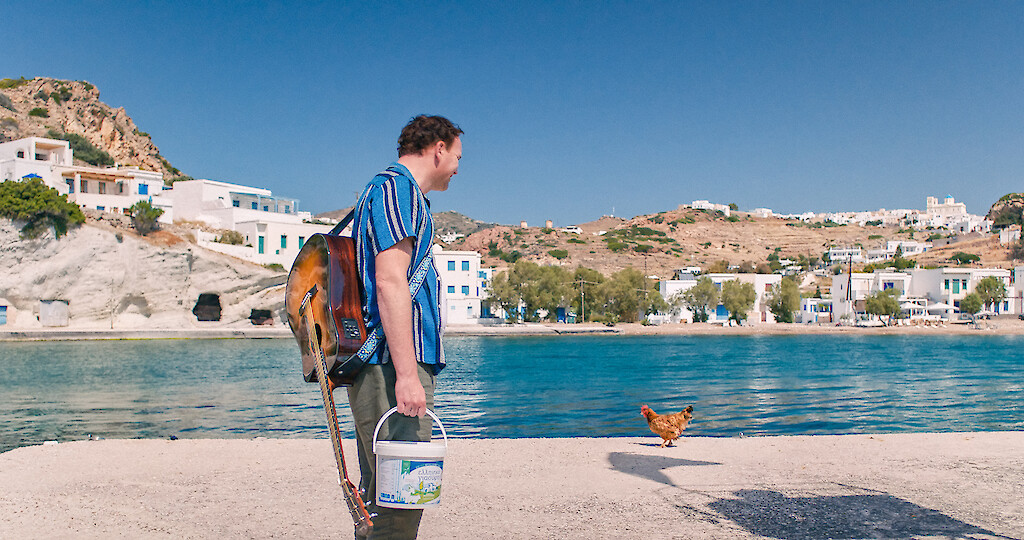 Johannes (Thomas Stipsits), a guitar on his back and a bucket in his hand, looking at a chicken in front of the sea.