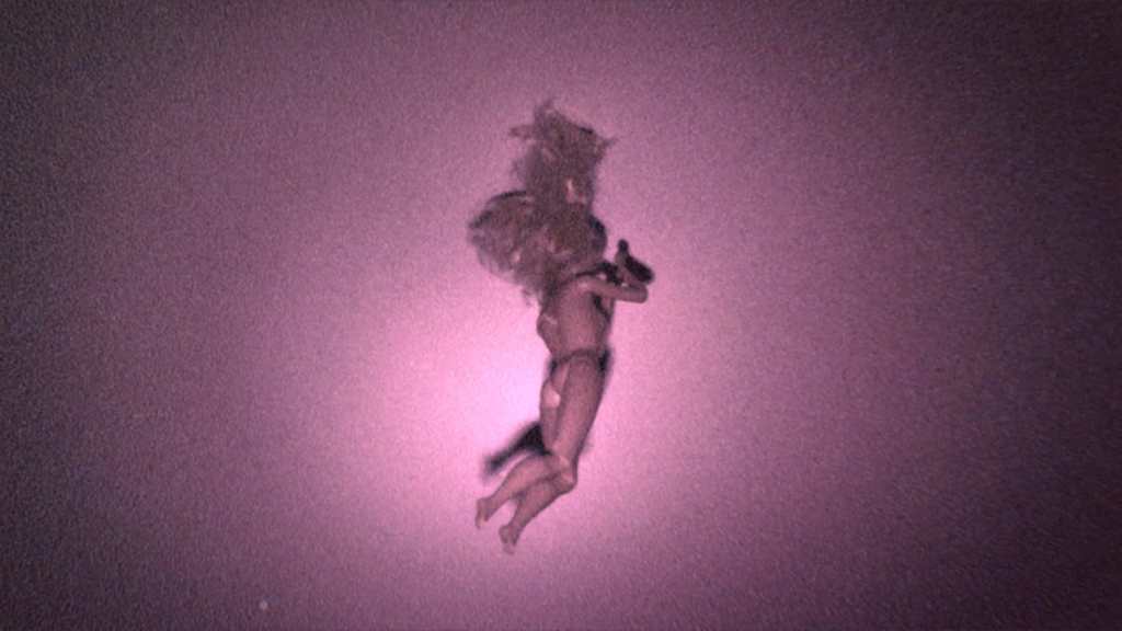 A barbie doll on a pink background. it seems to be floating just below the ceiling. The image is grainy.