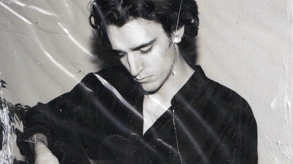 Tamino holding a guitar tht is just out of frame, looking down. The image is black and white with some white color over it that makes it look crumpled and old. 
