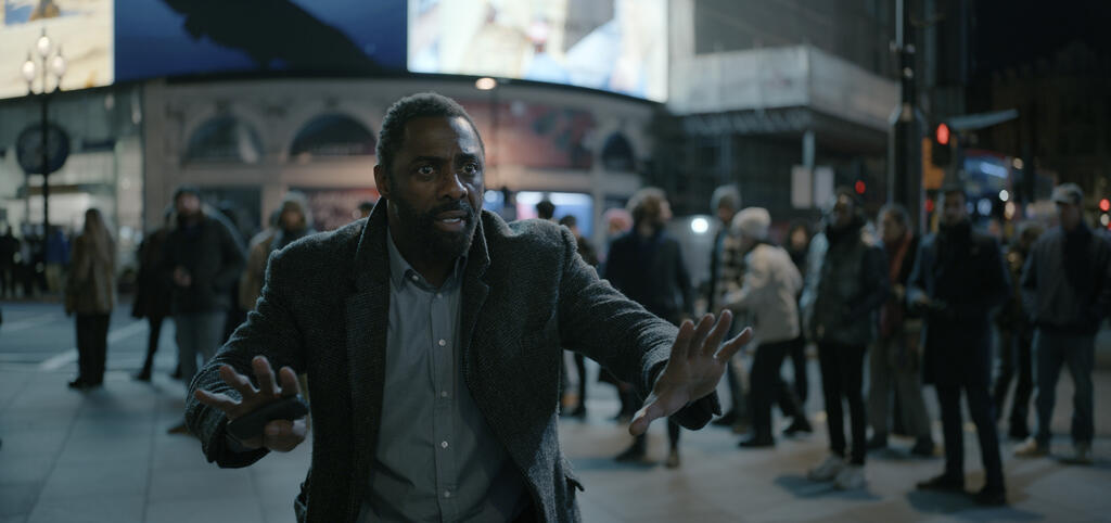 Luther (Idris Elba) standing on Piccadilly Square, making a calming gesture, a look of panic on his face.