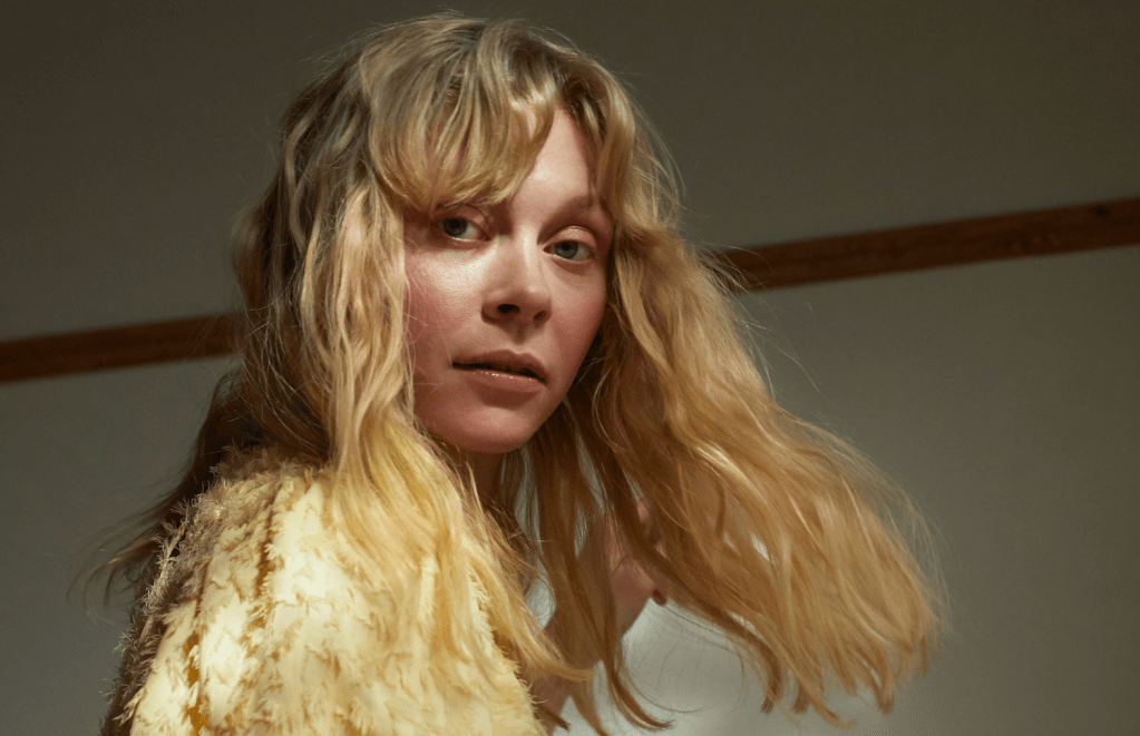 A young, white, blonde woman with long hair, looking over her shoulder at the camera, wearing a yellow top with feathers.