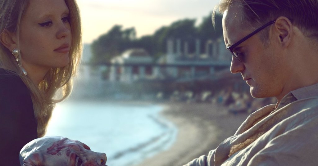 Gabi (Mia Goth) talking to James (Alexander Skarsgård) on the beach as he looks at a mask in his hands.