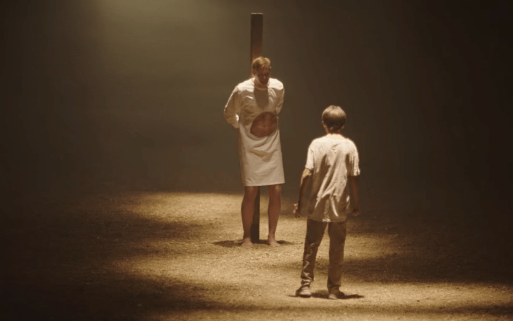 James (Alexander Skarsgård) tied to a pole in a torn white nightgown, a young boy standing before him.