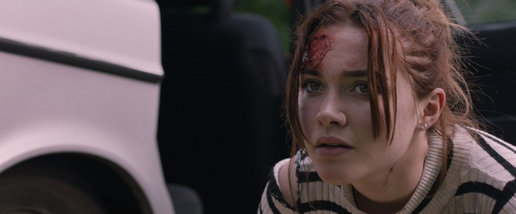 Angela (Florence Pugh) crouching next to a car with a wound on her forehead.
