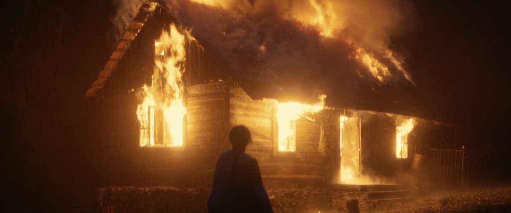 A burning hut, the shadow of a presumably female figure in front of it.