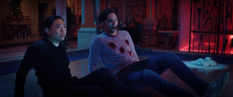 Rebecca (Awkwafina) and Renfield (Nicholas Hoult) sitting on the floor. He is covered in blood and bullet holes.