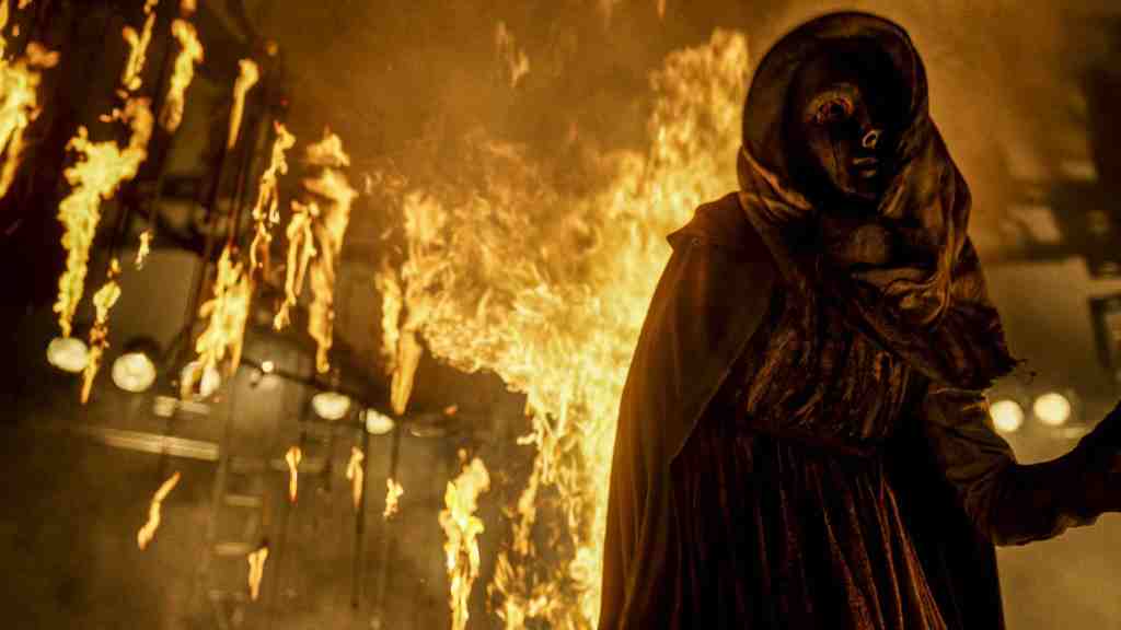 A masked and robed female figure in front of a fire. 