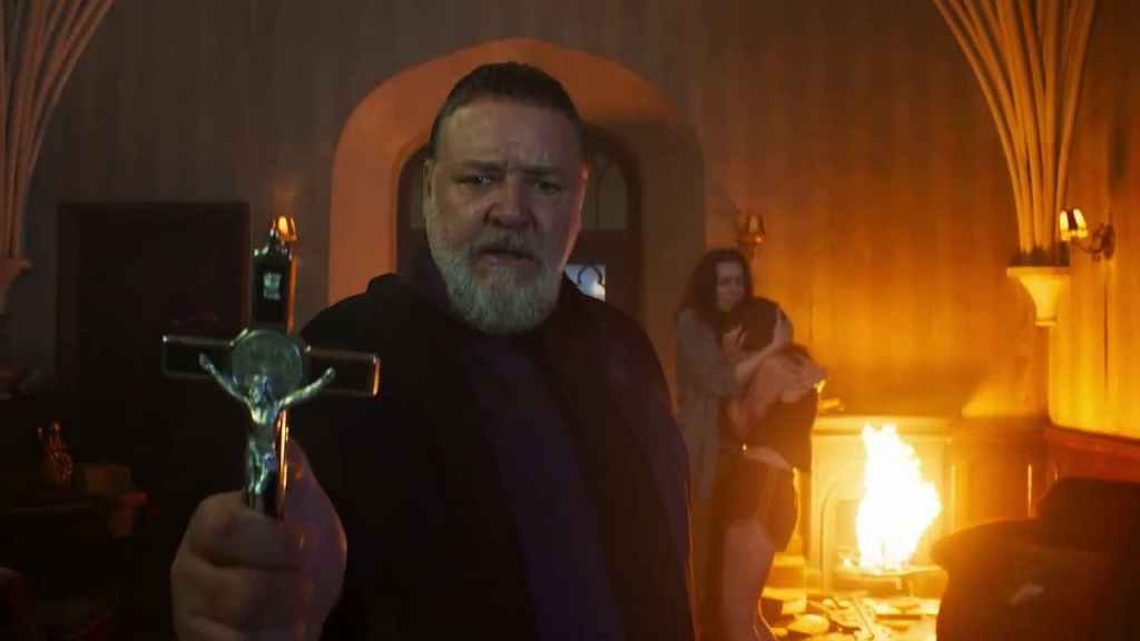 Gabriele Amorth (Russell Crowe) holding out a cross. Behind him, the room is on fire and we can see two women hugging each other.