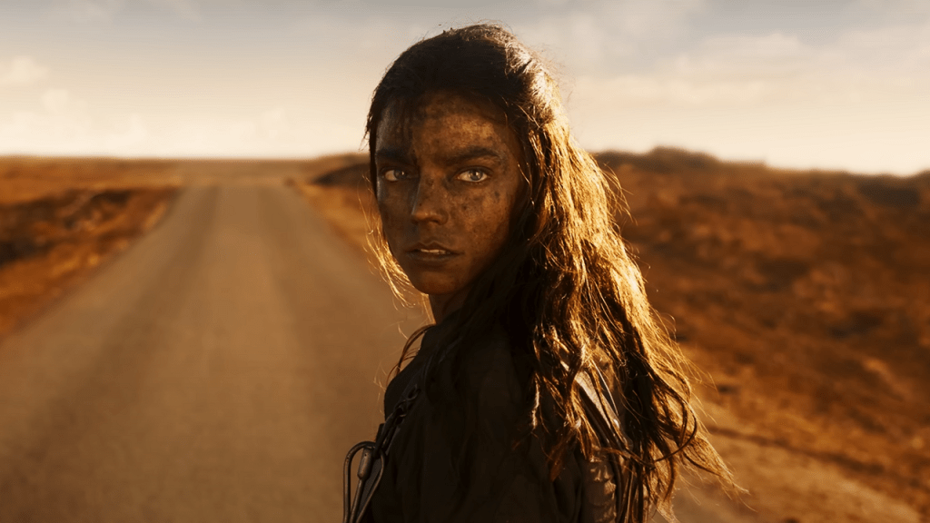 A disheveled and dirty Furiosa (Anya Taylor-Joy) on a road in the desert, looking back over her shoulder.