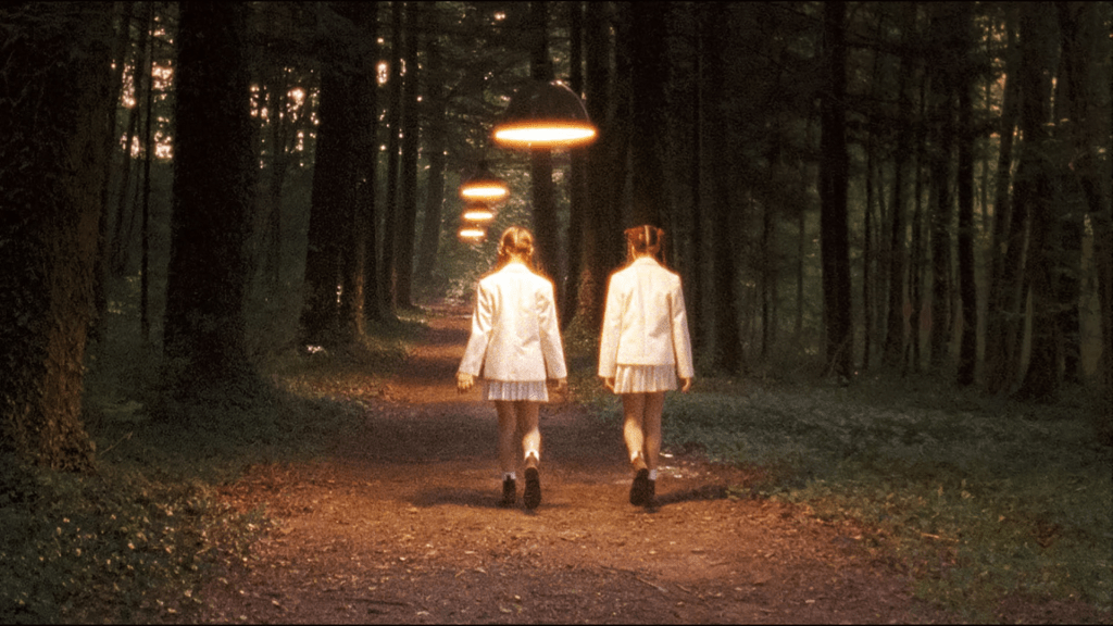 Two girls seen from behind as they walk trhough a forest path lit by lamps hanging over their heads.