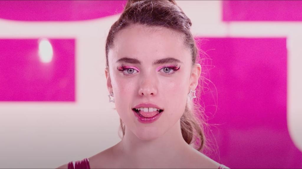 Sue (Margaret Qualley) in colorful make-up, looking straight at the camera with a smile.