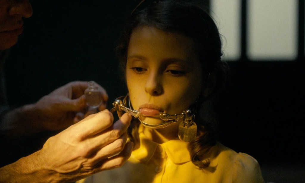 Mia (Romane Hemelaers) having her spit collected with a metal frame around her chin.