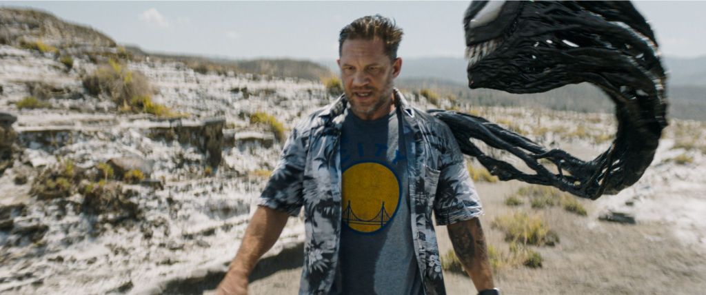 Eddie (Tom Hardy) walking through the desert, Venom's head emerging from his shoulder to look at him as they talk.