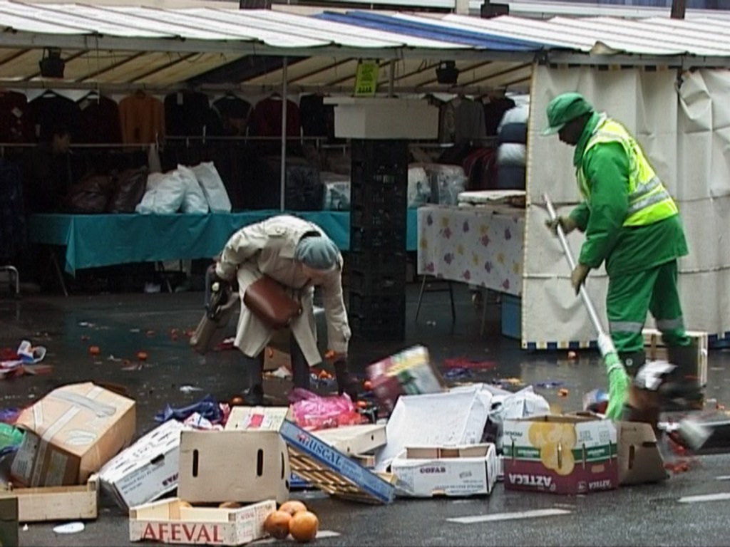 An older woman leaning over a few discarded boxes at a market while a cleaner sweeps them up.
