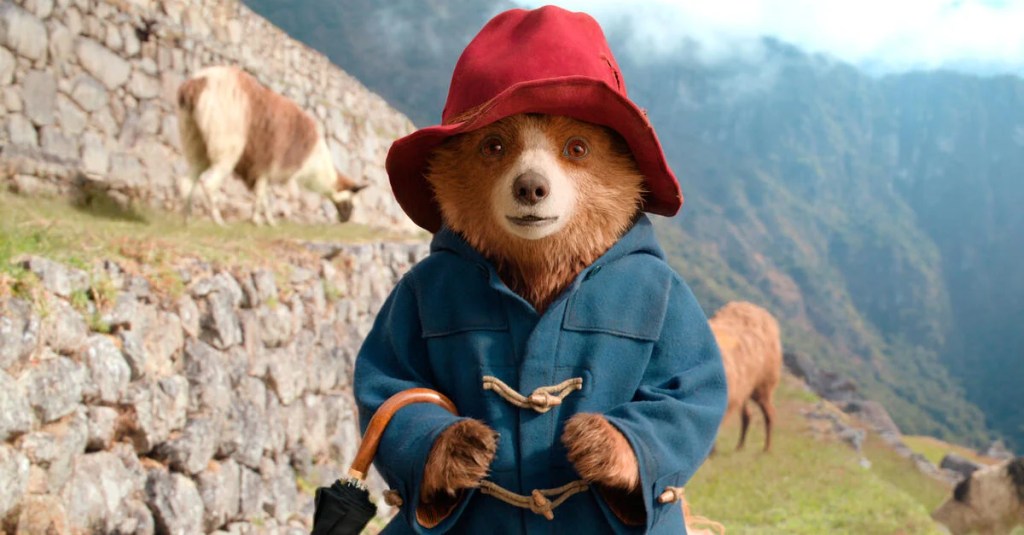 Paddington standing in some ruins in the mountains, llamas grazing behind him.