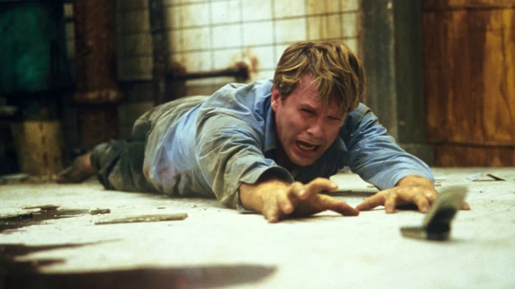 Lawrence Gordon (Cary Elwes) lying on the floor, desperately trying to get to a cell phone.