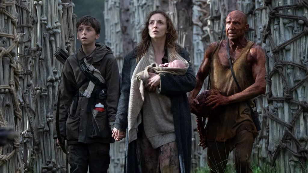 Spike (Alfie Williams), Isla (Jodie Comer) and Kelson (Ralph Fiennes) standing between towers made of bone. Isla is cradling a baby to her chest.