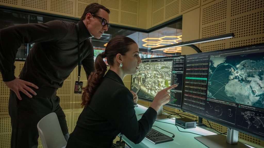 George (Michael Fassbender) looking at the computer screen of Clarissa (Marisa Abela) who is pointing at a world map.
