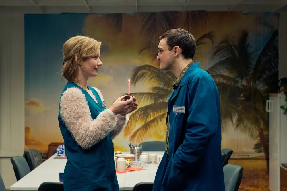 Christian (Franz Rogowski) and Marion (Sandra Hüller) in the employee break room. She is cradling a tiny cake with a single birthday candle. They are smiling softly at each other.