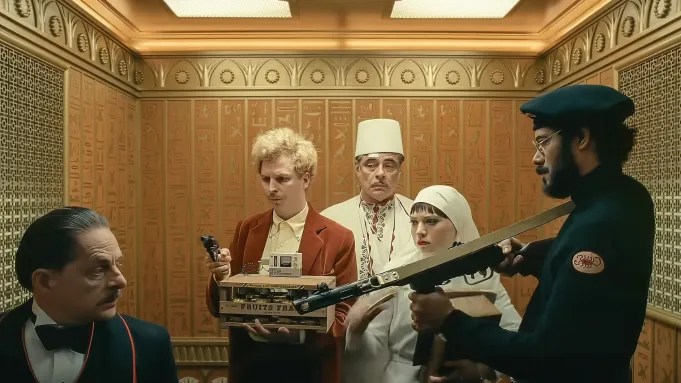 Zsa-zsa (Benicio Del Toro), Liesl (Mia Threapleton), Björn (Michael Cera) and Sergio (Richard Ayoade) all looking and partly pointing guns at the elevator porter. 