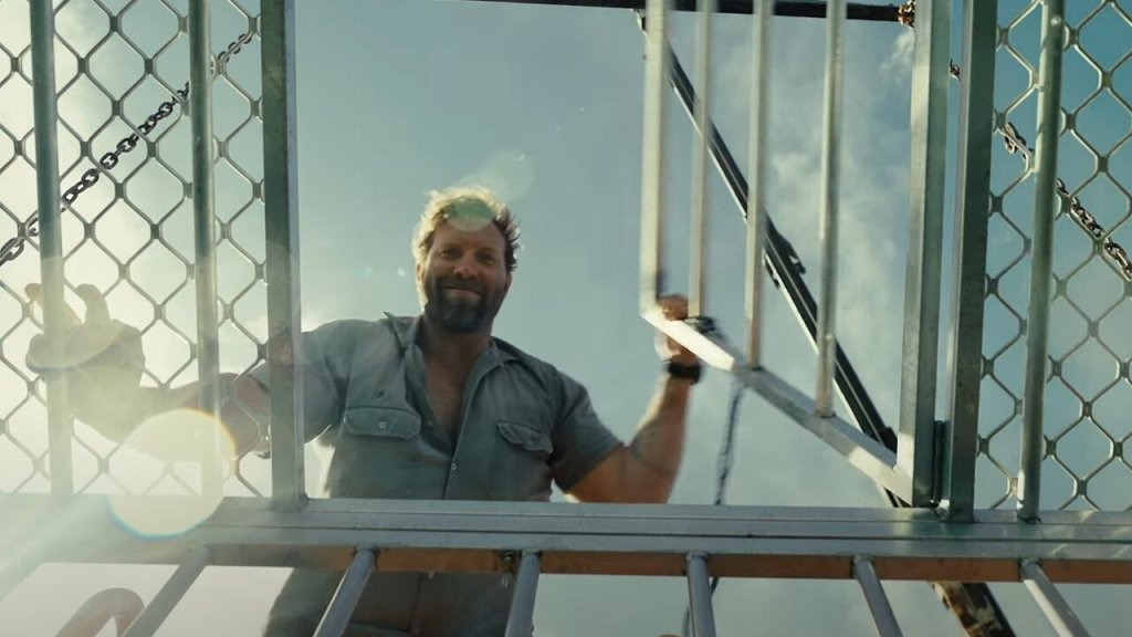 Tucker (Jai Courtney) looking down into a shark cage. The perspective is from inside the cage. He is smiling.