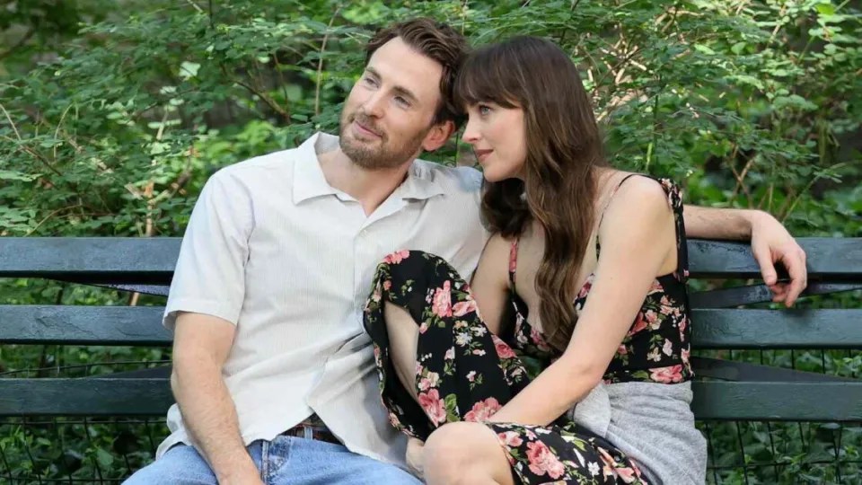 John (Chris Evans) and Lucy (Dakota Johnson) sitting on a parkbench, leaning against each other.
